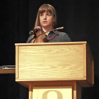 Claire Tonry stands at a wooden podium with a microphone, speaking during an award presentation at the University of Oregon. She wears a dar