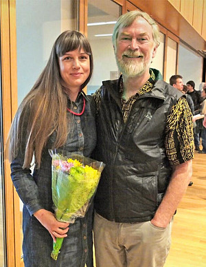 Claire Tonry and Charlie Tebbutt stand smiling together indoors; Claire holds a bouquet of flowers, and both are dressed in casual, professi