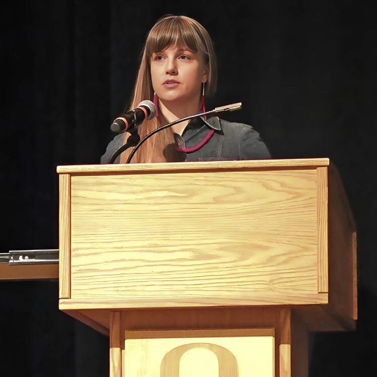 Claire Tonry stands at a wooden podium with a microphone, speaking during an award presentation at the University of Oregon. She wears a dar