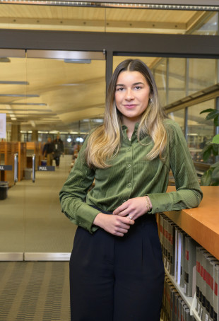Jeni looking at the camera inside the library. She is wearing a green button-up top and a black skirt.
